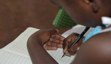 Lapsi kirjoittaa mustekynällä vihkoon, lähikuva käsistä ja kirjasta, taustalla sumea.