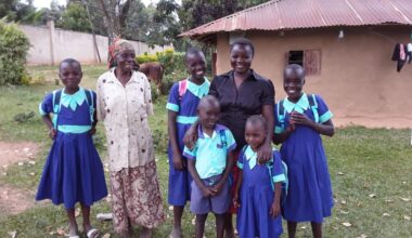 Ryhmä lapsia ja kaksi aikuista seisoo hymyillen pihalla savitalon edessä, koulupukuiset lapset selkärepulla.