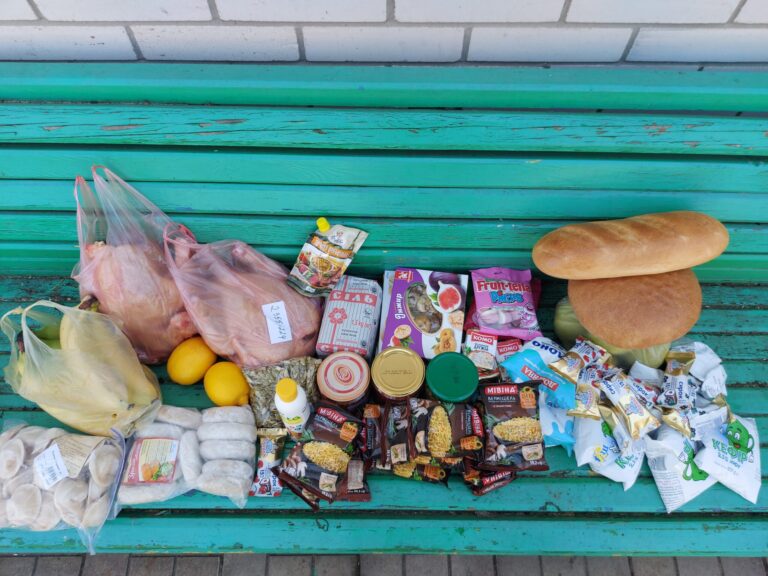Ruokatarvikkeita turkoosilla penkillä: leipää, lihaa, hedelmiä, paketteja ja muovipusseja, yhteensä runsas valikoima.