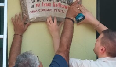 Kaksi henkilöä kiinnittää muistotaulua seinälle; vasemmalla porakone, oikealla kiinnittävät kädet.