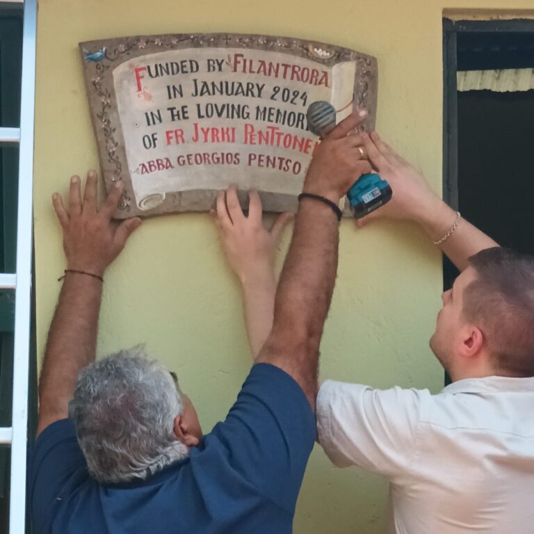 Kaksi henkilöä kiinnittää muistotaulua seinälle; vasemmalla porakone, oikealla kiinnittävät kädet.