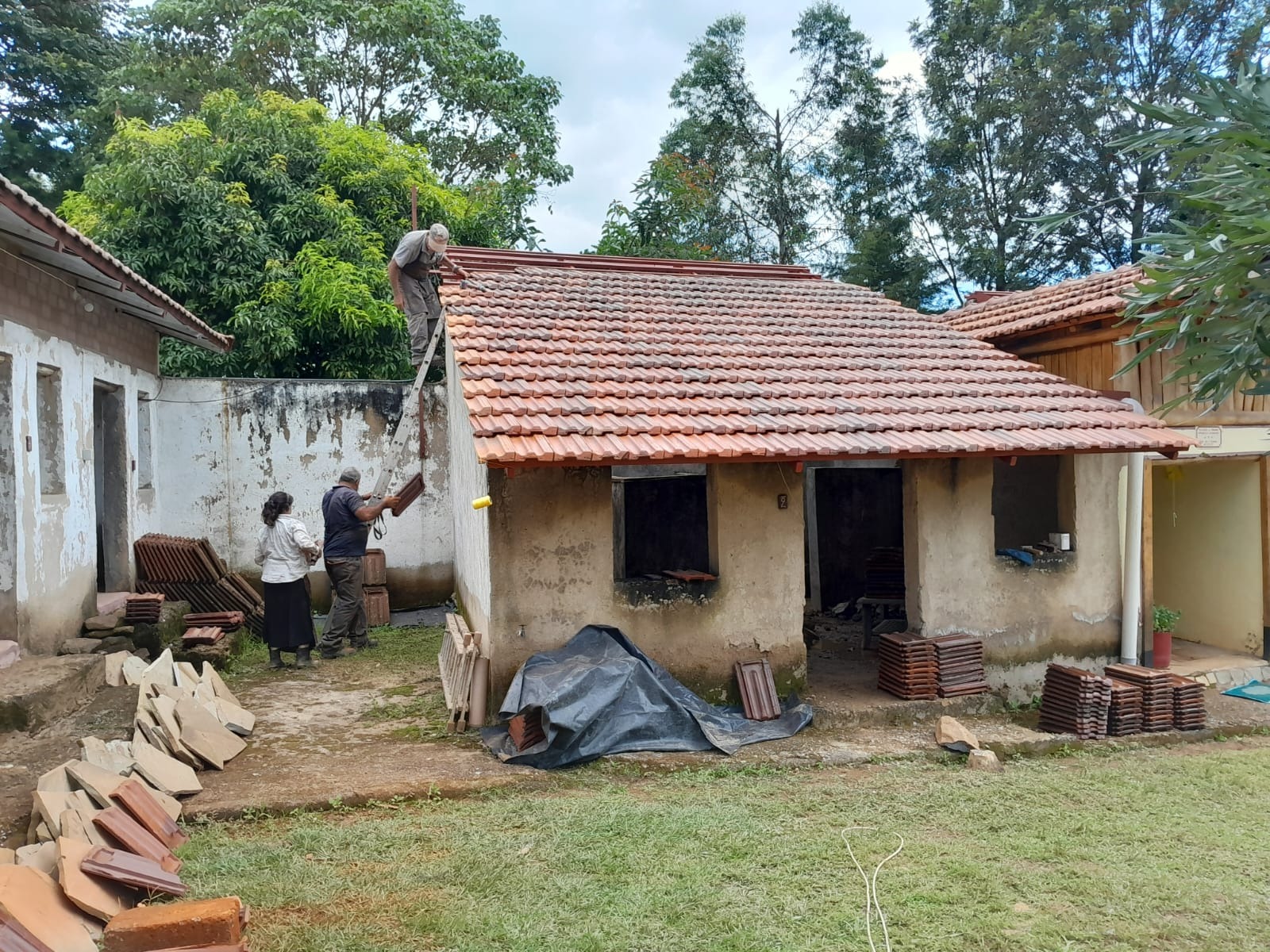 Kolme henkilöä korjaa pientä tiilikattoista taloa vehreän pihan ympäröimänä. Yksi henkilö on katolla.
