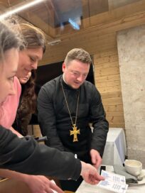 Kuvassa on pappi, joka jakaa papereita useille henkilöille, taustalla puupaneeliseinä.