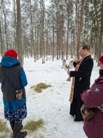 Lumiseen metsään kokoontunut ryhmä ihmisiä, joista yksi pitää kädessään esinettä ja toinen katsoo, taustalla poroja.