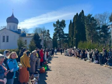Ihmiset seisovat jonossa kirkon edustalla kauniina kevätpäivänä.