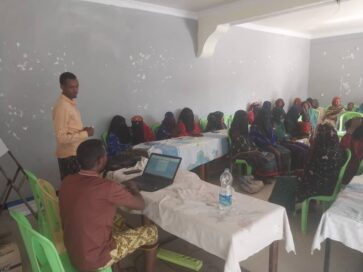 Ihmiset istuvat luokkahuoneessa muovituoleilla, yksi henkilö seisoo ja yksi istuu kannettavan tietokoneen ääressä.