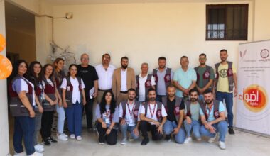 Ryhmä ihmisiä seisoo ja istuu sisätiloissa mainoskylttien edessä, useilla on yllään virkapuvut.