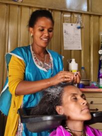 Nainen pesee asiakkaan hiuksia kampaamossa. Asiakas rentoutuu silmät suljettuina, kampaaja hymyilee.