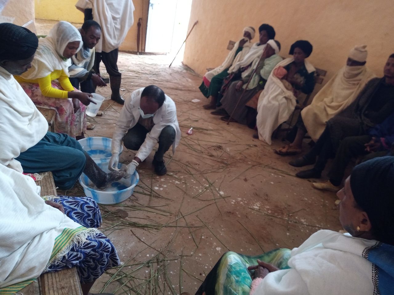 Hoitaja pesee potilaan jalkaa suuressa astiassa, muiden ihmisten istuessa penkillä taustalla.