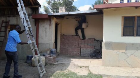 Kaksi henkilöä työskentelee talonrakennuksen parissa, toinen asettaa tikkaita, toinen poraa seinää.