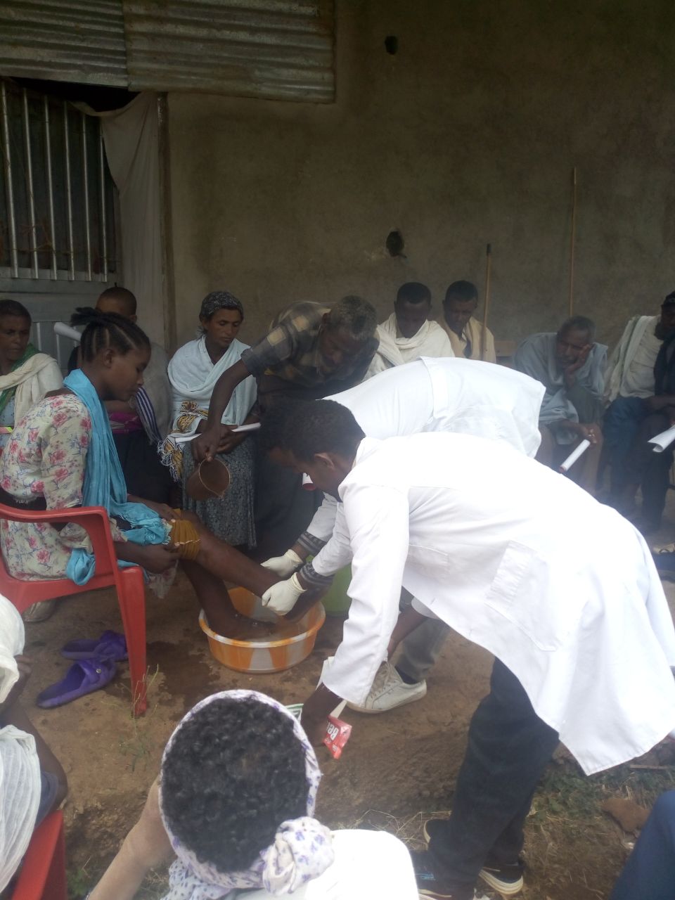 Henkilö pesee jalkoja metallikulhossa ulkona, muiden istuessa ympärillä. Tapahtuma vaikuttaa yhteisölliseltä.