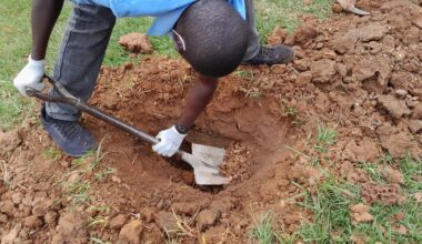 Henkilö kaivaa kuoppaa maahan lapion avulla. Hänellä on valkoiset hanskat ja sininen paita, ympärillä ruohoa.