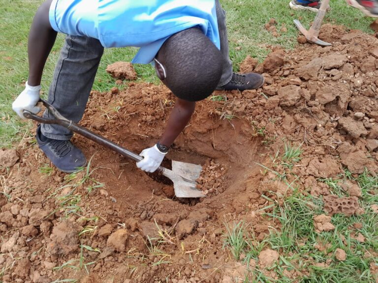 Henkilö kaivaa kuoppaa maahan lapion avulla. Hänellä on valkoiset hanskat ja sininen paita, ympärillä ruohoa.
