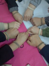 Kahdeksan kättä, jokaisella samanlainen punottu rannekoru, muodostavat ympyrän vaaleanpunaisen ja valkoisen kankaan päällä.