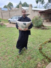 Mies seisoo mustassa kaavussa vihreällä nurmikolla pitäen suurta säkkiä sylissään. Taustalla auto ja rakennus.