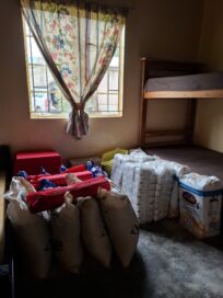 Pussit lattialla huoneessa, jossa on kerrossänky ja ikkuna kukkaverhoilla. Taustalla pinottuja tuotteita.