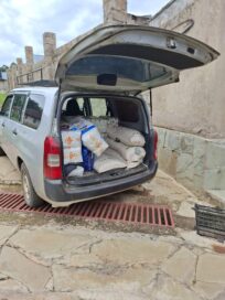 Auton takaluukku auki, täynnä suuria jauhosäkkejä. Auto seisoo kivetyksellä rakennuksen vieressä.