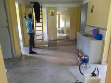 Mies katselee tikkaita käytävällä; toinen henkilö nousee tikkailla. Seinillä kuvia, pesuallas oikealla.