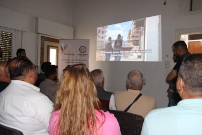 Ihmiset istuvat huoneessa katsomassa projektoria, jossa kuvataan rakennusvaurioita. Kankaalla tekstitys englanniksi.