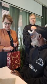 Nainen pitää laukusta kiinni, vieressä vauvaa pitelevä henkilö ja toinen istuva henkilö, jolla on musta huppari.