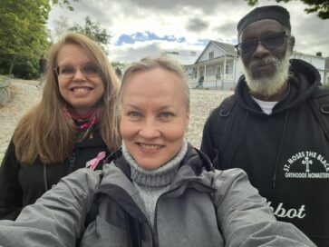 Kolme ihmistä poseeraa ulkona edessä hirsitalon. Taustalla pilvinen taivas ja vehreitä puita.