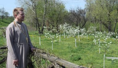 Mies seisoo aidan edessä keväisessä puutarhassa, jossa on kukkivia puita. Hän käyttää pitkää kaapua ja kultaista kaulakorua.