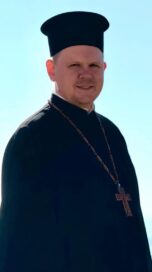 Mies tumma puku ja hattu yllään, kaulassa ketju ja risti. Taustalla sininen taivas.