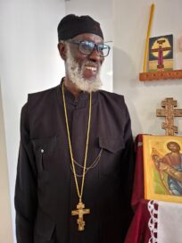 Mies mustassa asussa, kultainen risti kaulassa, hymyilee maalauksen ja ristin vieressä kirkollisessa ympäristössä.