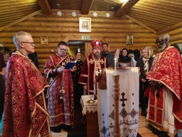 Ortodoksinen kirkonmeno hirsirakenteisessa kappelissa. Piispa ja papit punaisissa kaavuissa, pöydällä palava kynttilä.