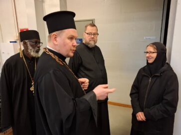 Neljä henkilöä keskustelee sisätiloissa; kaikki pukeutuneet tummiin uskonnollisiin asuihin ja hattuihin.