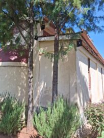 Punakattoinen rakennus puiden ja pensasaidan keskellä kirkkaassa päivänpaisteessa.