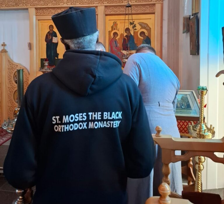 Mies ortodoksikirkossa, jossa on ikoneja ja koristeellisia puuleikkauksia. Hupparissa lukee St. Moses the Black.