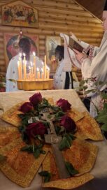 Ortodoksinen seremonia, jossa kynttilöitä, ruusuja ja krusifiksi. Taustalla ikoneita ja pappi lukee tekstikirjaa.