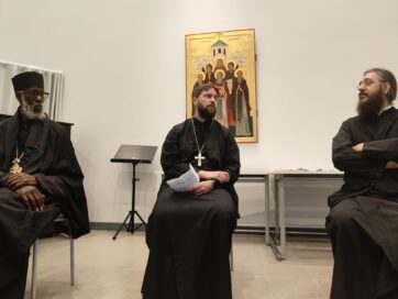 Kolme pappia mustissa kaavuissa istumassa kirkollisessa tilassa, taustalla ikoni.