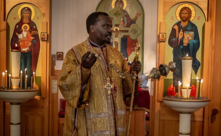 Pappi suitsuttaa kirkkosalissa ikonien edessä, ympärillä kulttuurisesti rikkaasti koristellut seinät ja kynttilät.