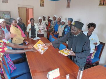 Ryhmä ihmisiä kokoontuneena huoneeseen, pappi ojentaa pyhän kuvan toiselle henkilölle.