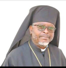 Mies mustassa papin kaavussa ja päähineessä, silmälasit, helminauha kaulassa, valkoinen tausta.