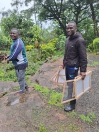 Kaksi miestä seisoo ulkona vihreässä ympäristössä, toinen heistä pitää kädessään mehiläistarvikkeita.