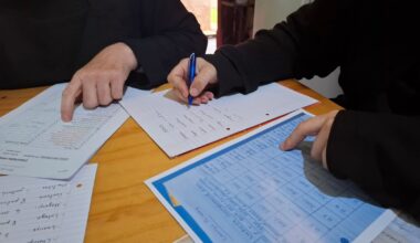 Kaksi henkilöä tarkistaa ja kirjoittaa muistiinpanoja kirjanpitoon pöydän ääressä.
