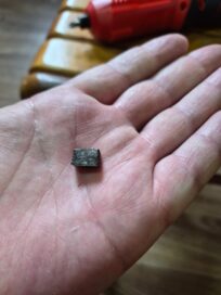 Käsi, jossa pidetään pientä mustaa kuutiomaista esinettä, taustalla näkyy punainen työkalu.